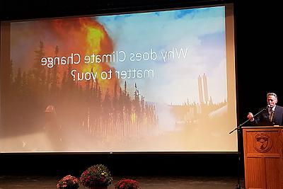 Image of former Vermont Governor Peter Shumlin standing at a podium in the Greenhoe Theater. A screen projection shows an image of a forest fire and text reading Why Does Climate Change Matter to You? 