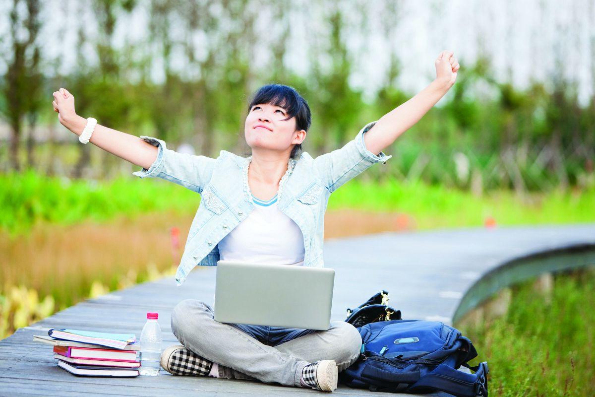 Female student sitting cross-legged outside with open laptop on her lap. She is stretching her arms up in the air and looking up at the sky.